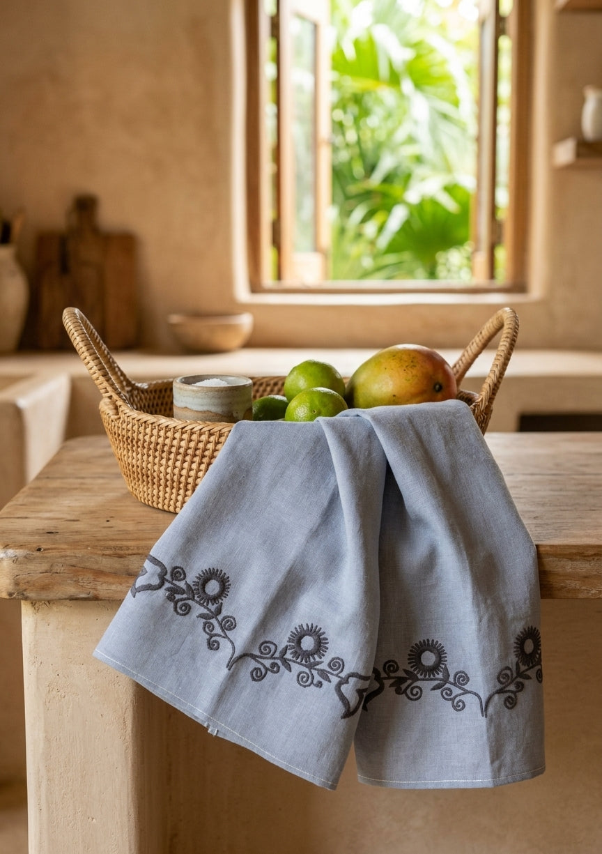 Soleil Stitch Dish Towel – soft blue chambray with navy sunflower border embroidery, styled with fruit basket