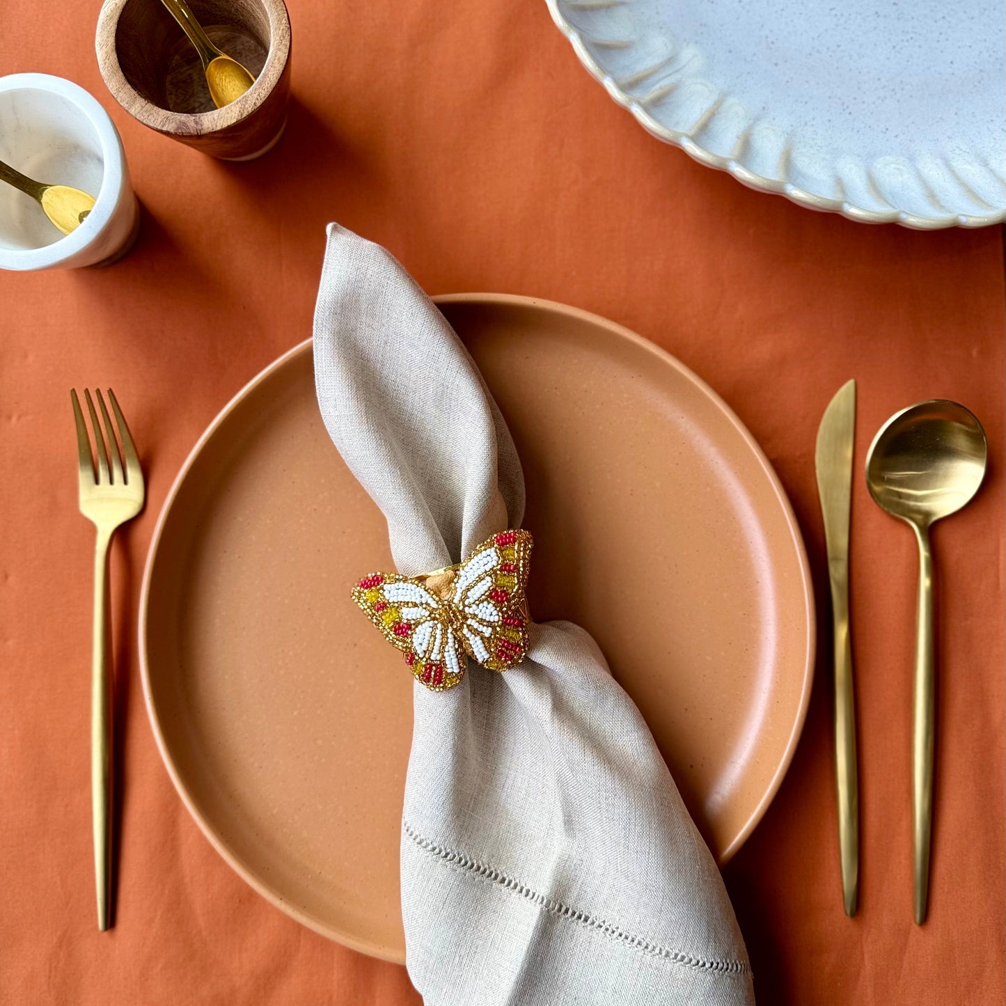 Dining table setting with beige plate, gold fork and knife, and butterfly napkin ring on a terracotta surface.